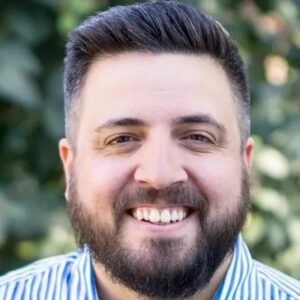 Close-up portrait of SEO consultant and content marketer AJ Ghergich smiling with a full beard in a blue-and-white striped shirt outdoors.