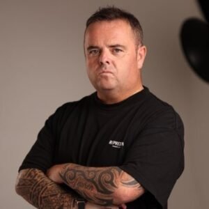 Portrait of Scottish SEO expert Craig Campbell with folded, heavily tattooed arms, wearing a black t-shirt against a studio background.