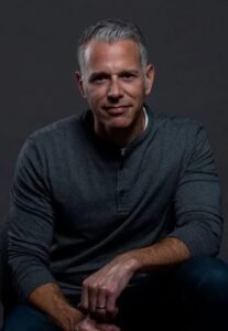 Portrait of B2B marketing strategist and author Lee Odden seated in a charcoal henley shirt against a dark studio background.