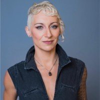 Portrait of SEO strategist and speaker Lily Ray with short blonde styled hair wearing a dark denim vest against a solid blue backdrop.