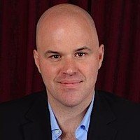 Headshot portrait of Search Engine Journal founder Loren Baker smiling in a black suit jacket and light blue collared shirt against a dark curtain backdrop.