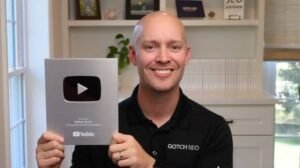 Nathan Gotch smiling while holding his YouTube Silver Creator Award in a black Gotch SEO polo shirt inside a home office.