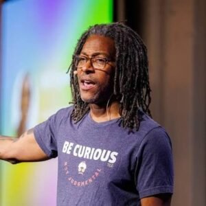 Digital marketing leader and Seer Interactive founder Wil Reynolds speaking on stage wearing glasses, dreadlocks, and a graphic t-shirt reading Be Curious Not Judgmental.
